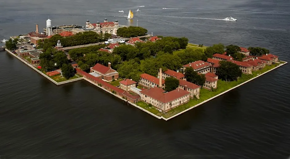 Color aerial photograph of Ellis Island today from the same angle: the same buildings with red roofs, now surrounded by mature trees and lawns