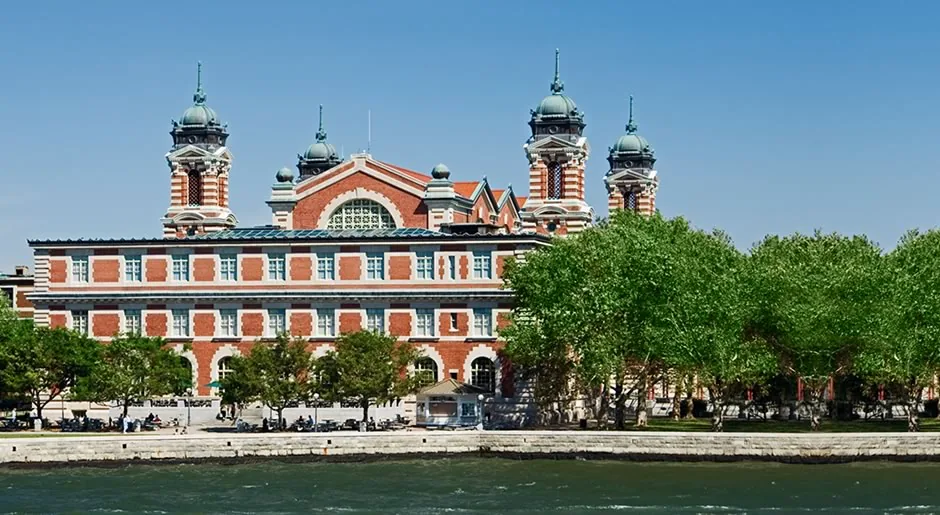 The restored main building today under a clear blue sky: red brick, white limestone trim and four copper-domed towers behind a line of green trees