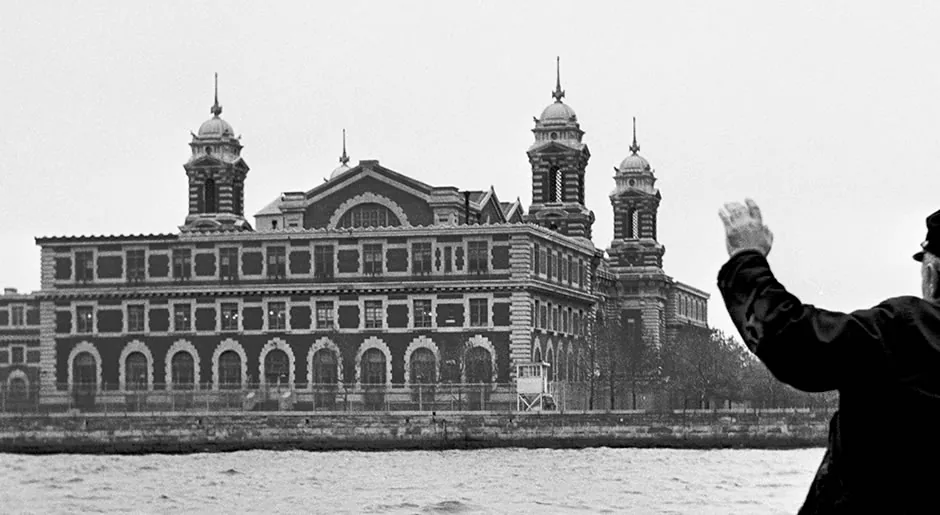 Black-and-white photograph of the Ellis Island main building seen from the water, a man in a cap raising his hand toward it in the foreground