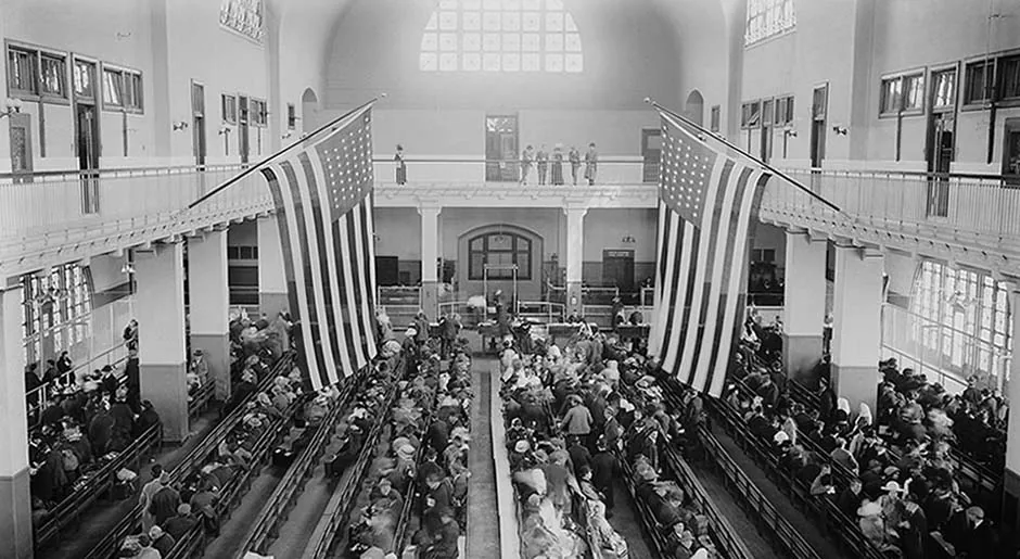 Black-and-white photograph of the Registry Room filled with immigrants seated on rows of benches, two large American flags hanging from the balcony
